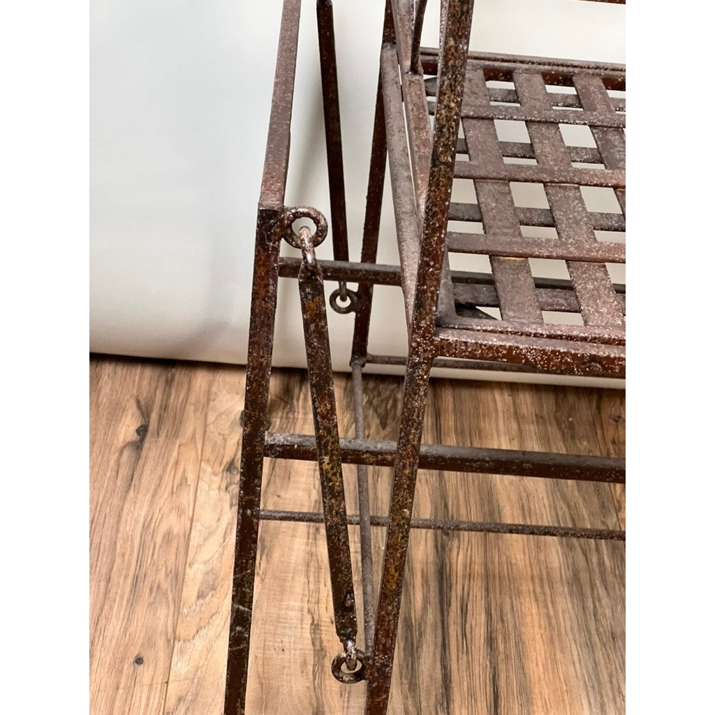 Close-up of a Vintage Regency Style Wrought Iron Porch Glider with a square grid seat, showing visible rust and wear on its metal frame. Set on a wooden floor with part of a plain white background visible. Brand: Unknown.
