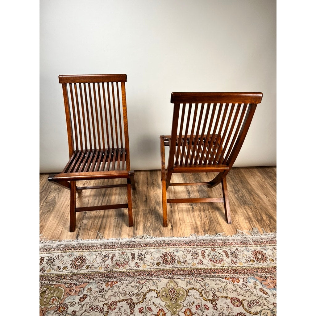 A pair of vintage mid-century modern style teak folding chairs are set on a wooden floor—one facing forward, the other angled. A patterned rug lies in front, complementing the plain wall in the background. Brand unknown.