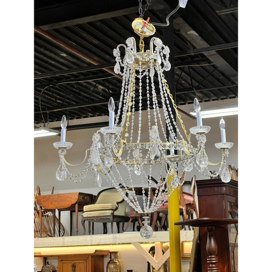 A Vintage Empire Style Brass and Rock Crystal Six Light Chandelier by Pearl City Antiques, featuring candle-shaped lights and draped bead strands, hangs in a store filled with antique furniture such as tables and chairs visible in the background.