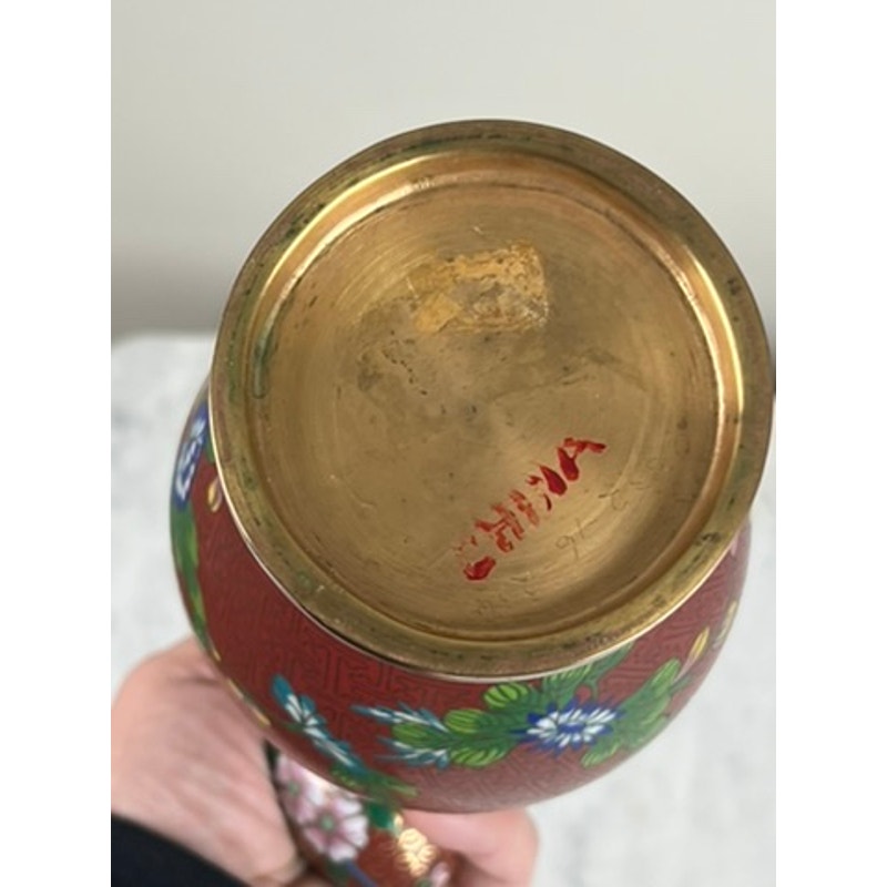 A close-up of the brass base of a Vintage Chinese Cloisonné Vase and Egg, held in hand. The vase features enameling with a red background, green leaves, and blue-and-white flowers, plus a red mark on the base. Brand unknown.