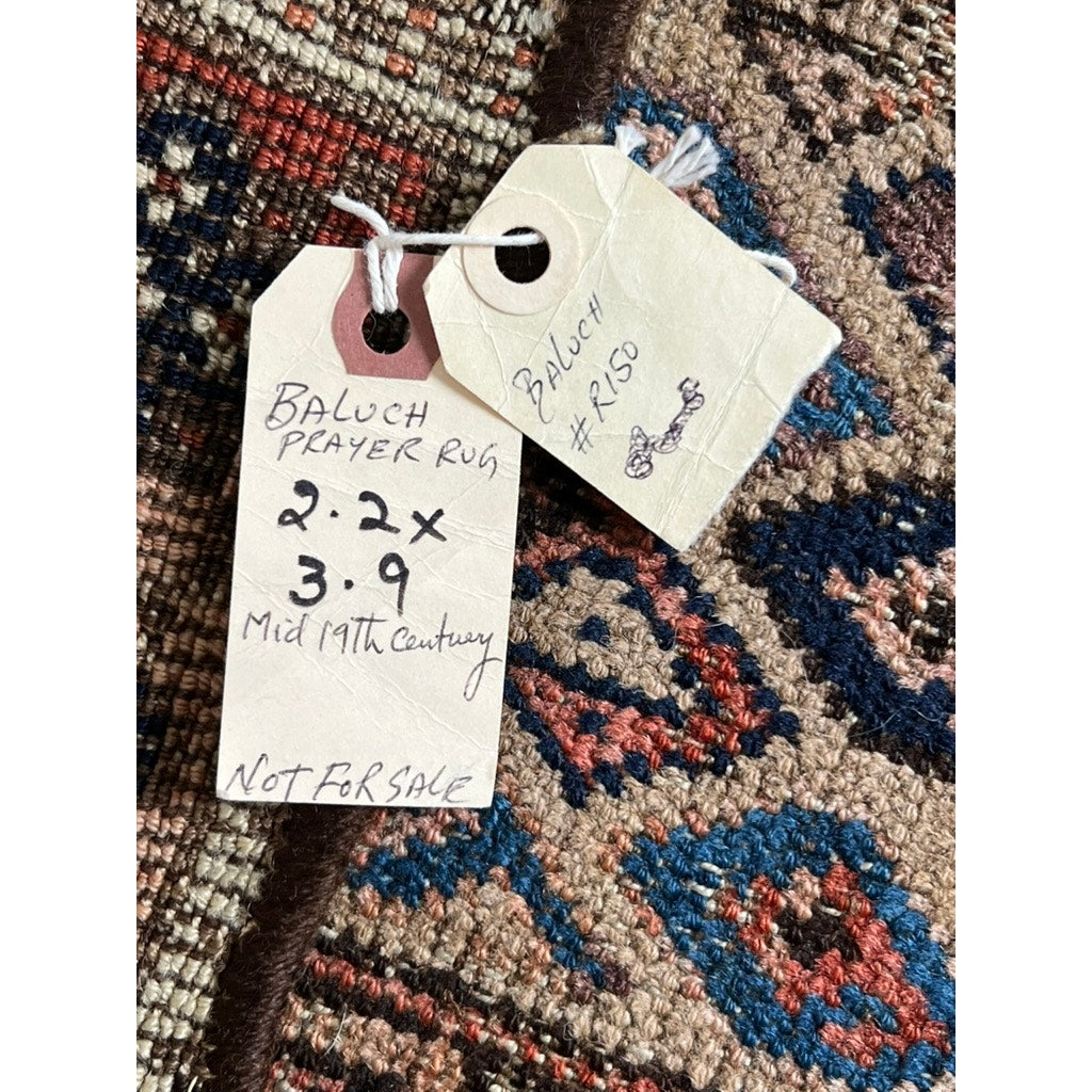 Two paper tags on a patterned antique rug: one reads “Rare Antique Baluch Prayer Rug with Diamond Lattice Field, Northeast Persia, c. Mid 19th Century – 2’2 x 3’9, NOT FOR SALE.” The other says “BALUCH #R150” and features a small drawing.