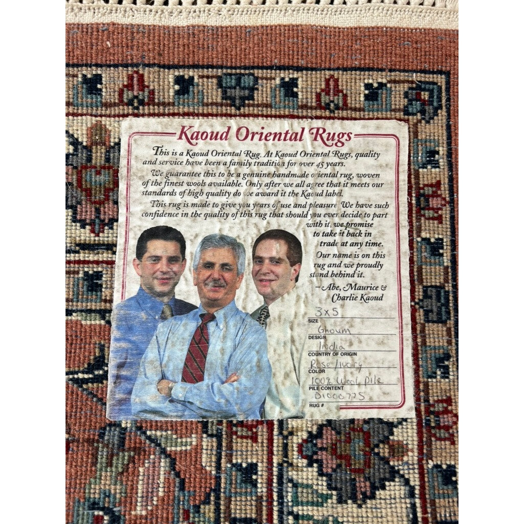 A label on the Vintage Oriental Indian Ghoum Hand Knotted Wool Rug 3x5’4 by Kaoud shows three men in dress shirts and ties, smiling together. It highlights the brand's quality and fine Indian hand-knotted wool craftsmanship.