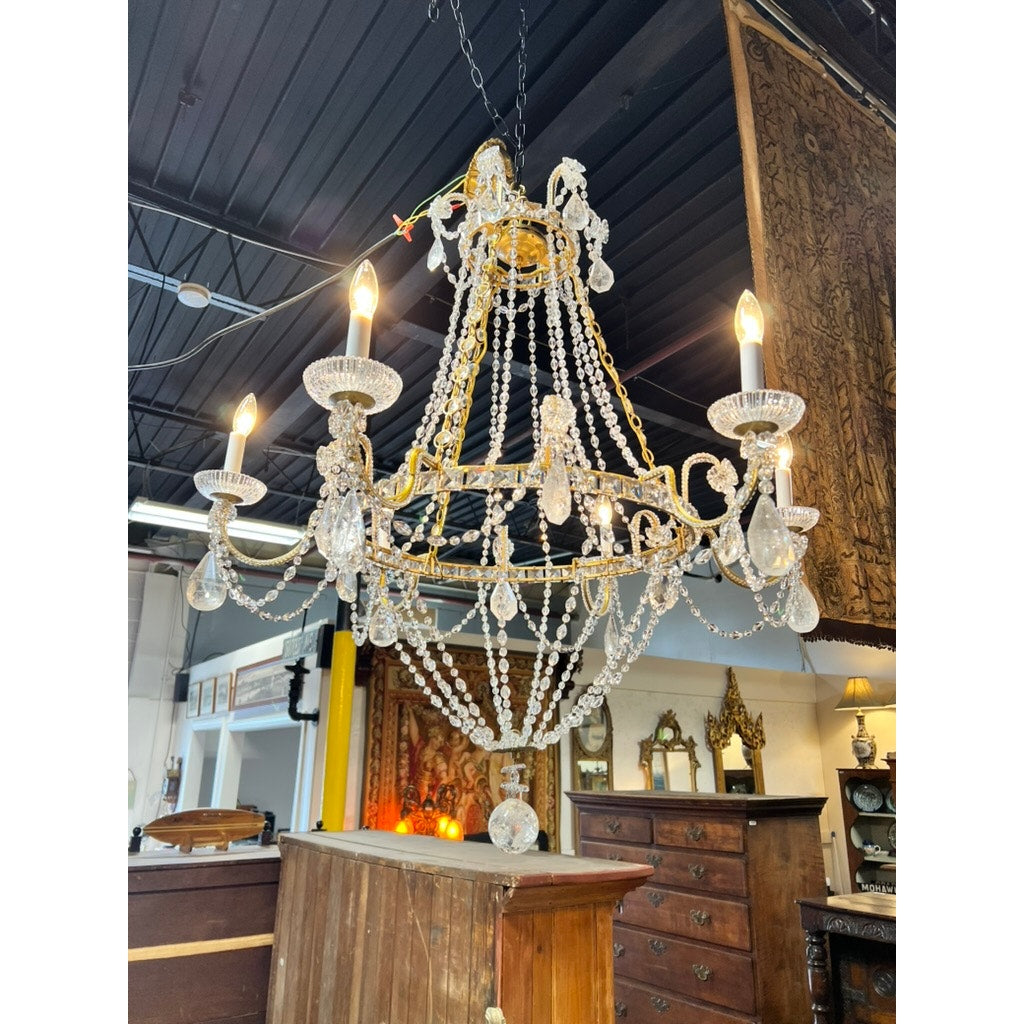 A Vintage Empire Style Brass and Rock Crystal Six Light Chandelier hangs from a dark ceiling in an antique store filled with wooden furniture and ornate mirrors. Brand: Unknown.