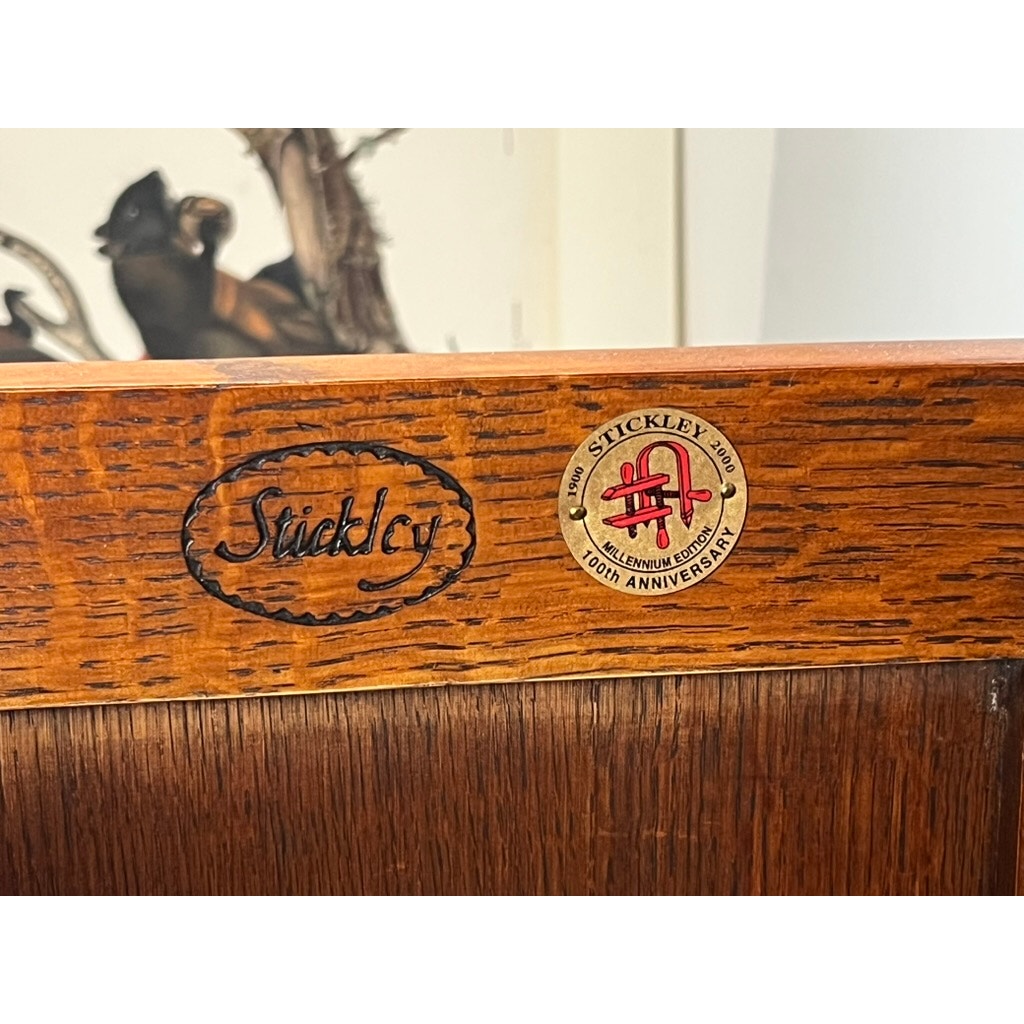 Close-up of a Vintage Stickley Harvey Ellis Millennium Edition Oak Blanket Chest, showing the oval Stickley logo on the left and a round 120th Anniversary sticker on the right—classic Arts & Crafts craftsmanship by Stickley.
