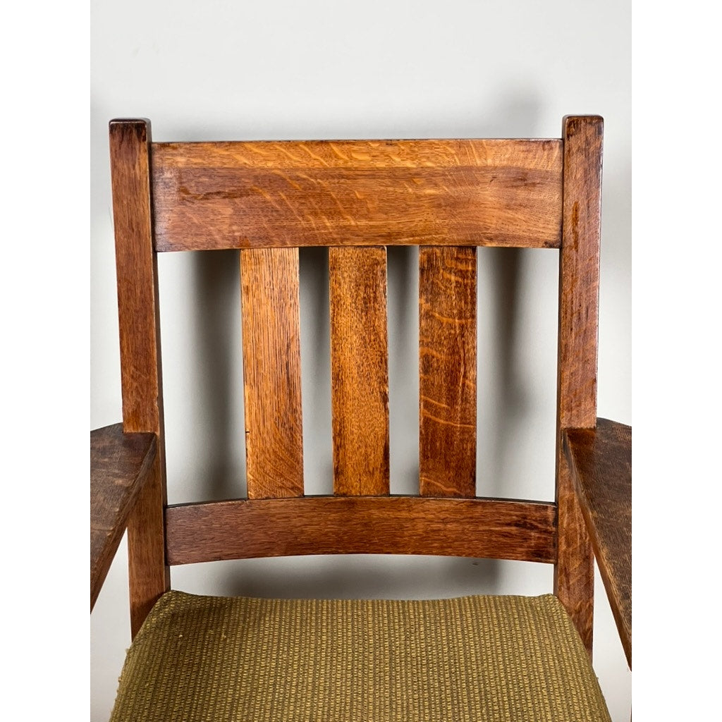 A close-up of a c. 1920 Antique Quarter Sawn Mission Oak Arm Chair by Quaint Furniture shows vertical slats, armrests, a textured light brown upholstered seat, and warm medium-tone wood with visible grain.