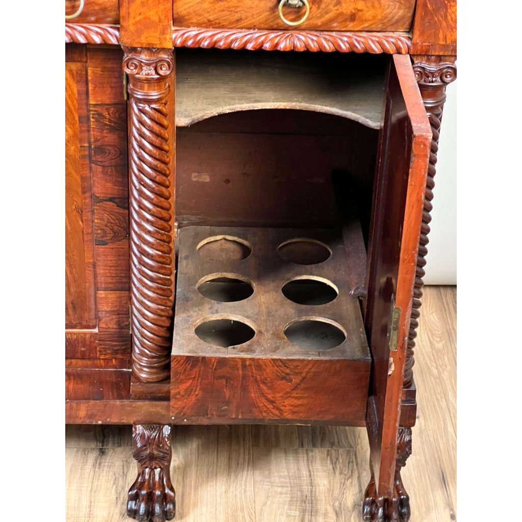 An Antique American Empire Mahogany with Rosewood Edge Banding Sideboard, likely Philadelphia c. 1840, stands on a wood floor. Ornate carving and claw feet adorn it; one door opens to reveal a shelf with four round bottle cutouts. Brand unknown.