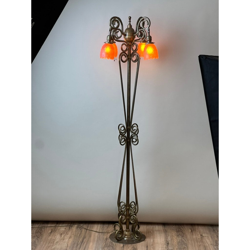 An Antique Art Nouveau brass floor lamp, attributed to Jules Cayette and Charles Schneider c. 1920, features ornate scrollwork and three glowing orange glass shades; it stands on a wooden floor against a plain light background.
