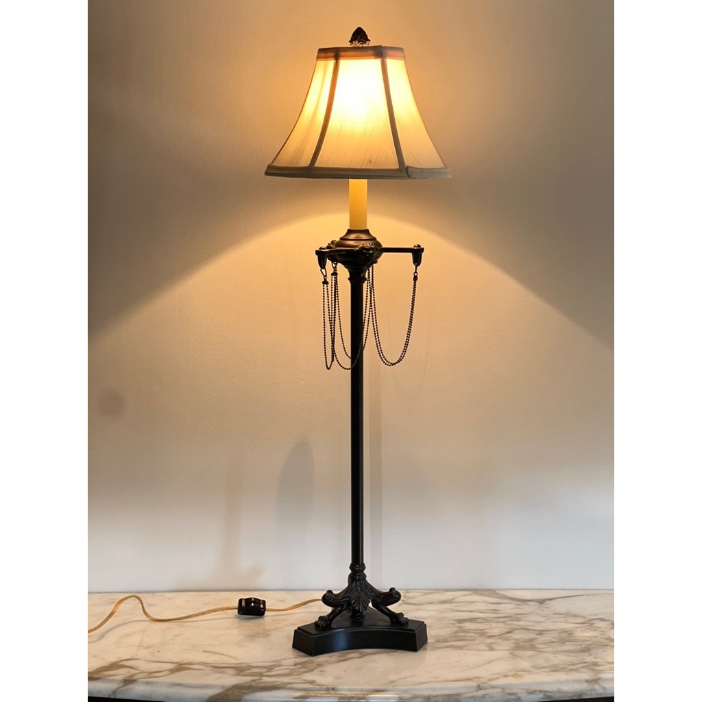 A Vintage French Empire Revival Table Lamp in oiled bronze finish with a beige shade sits lit on a marble surface. Decorative chains hang at its sides, and the power cord with switch lies across the tabletop. Brand unknown.