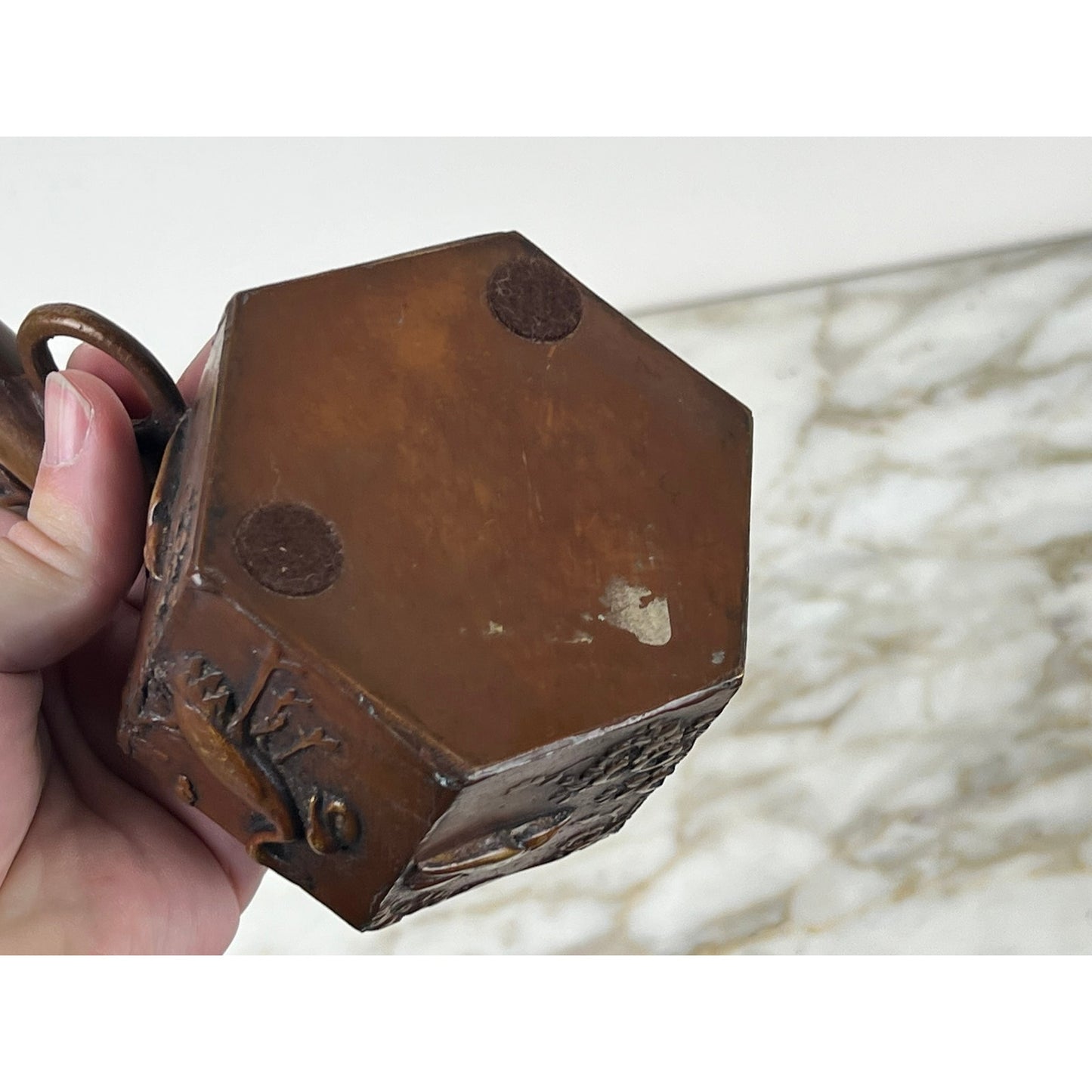 A hand holds an Antique Meiji Period Japanese Bronze Yatate Inkwell, hexagonal and brown with two round imprints; the background is a softly blurred, marbled surface in light tones. Brand is unknown.