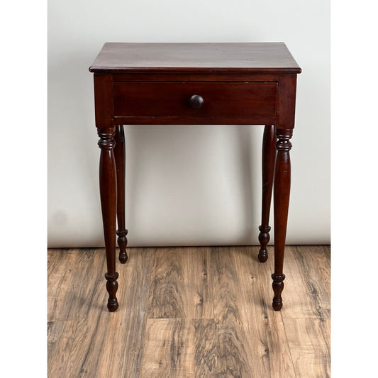 This Antique Sheraton Cherry One Drawer Stand, c. Mid 19th Century by Pearl City Antiques, features a single drawer with a round knob and four finely turned legs, beautifully showcased on a wood floor against a plain white wall.