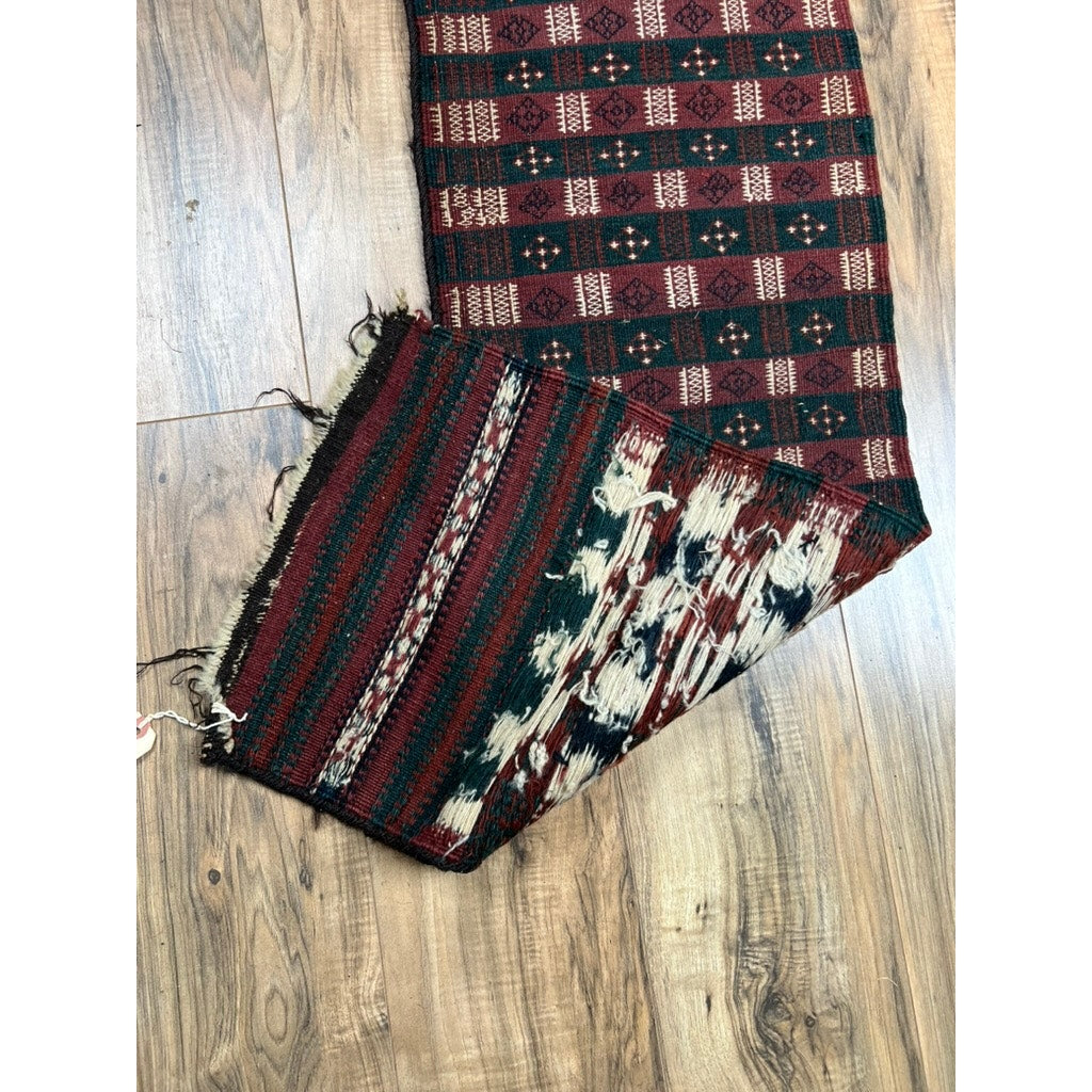An Antique Baluch Tribal Flatweave Sofreh Runner (c. Late 19th Century, 1'2 x 7'10) with red, black, and cream patterns rests on a wooden floor; its frayed bottom corner is partially folded, exposing intricate designs and loose threads.