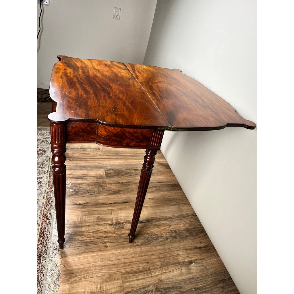 An Antique Federal Period Sheraton Cookie Corner Game Table c. 1830 with ornate legs stands on a hardwood floor by a white wall, one side extended to reveal its glossy finish and rich wood grain. Brand: Unknown.