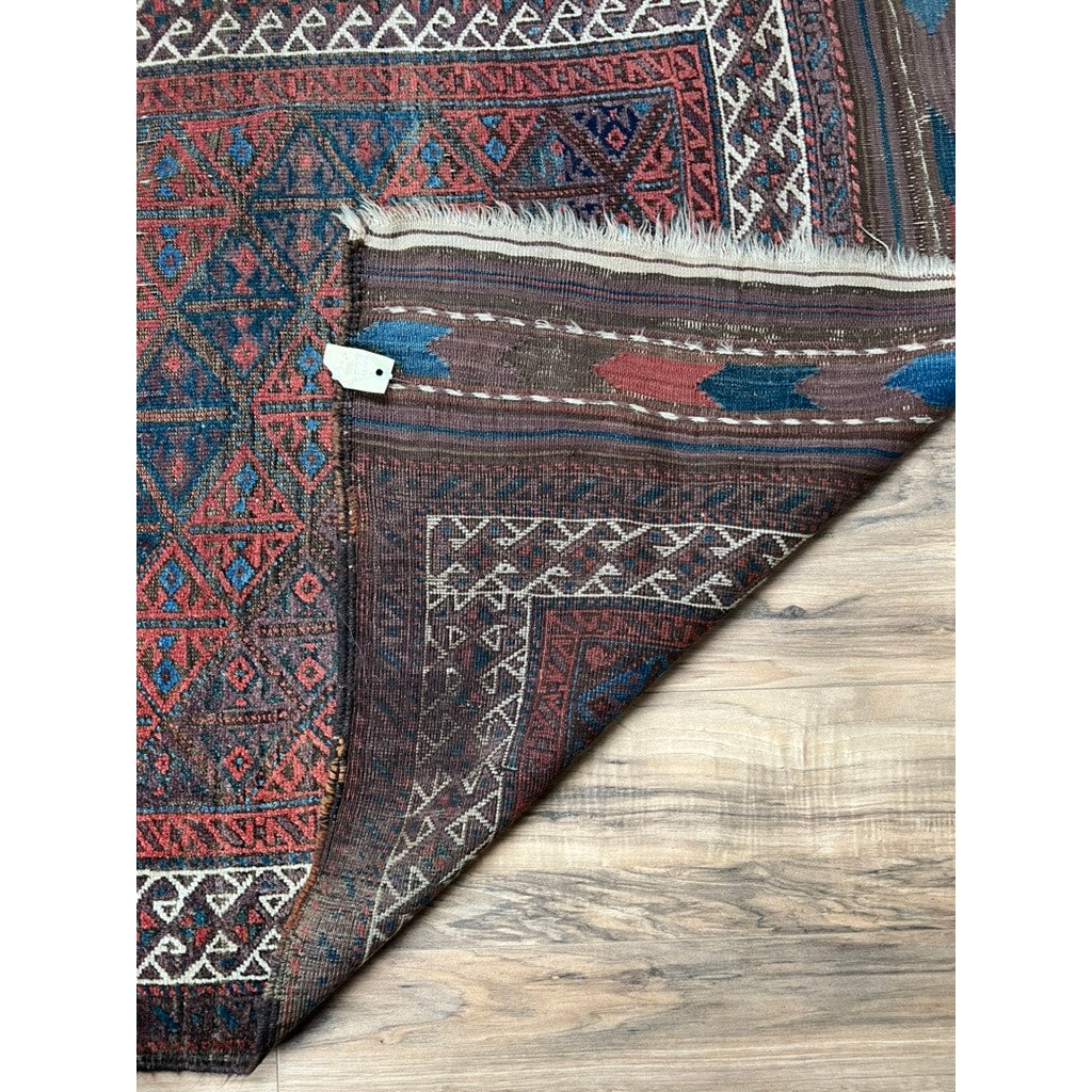 An Antique Baluch Rug with diagonal lattice and tree motifs (NE Persia, late 19th c., 3’3 x 6’) is partially flipped on a wooden floor, revealing its underside and fringed edge. Brand: None/Unknown.