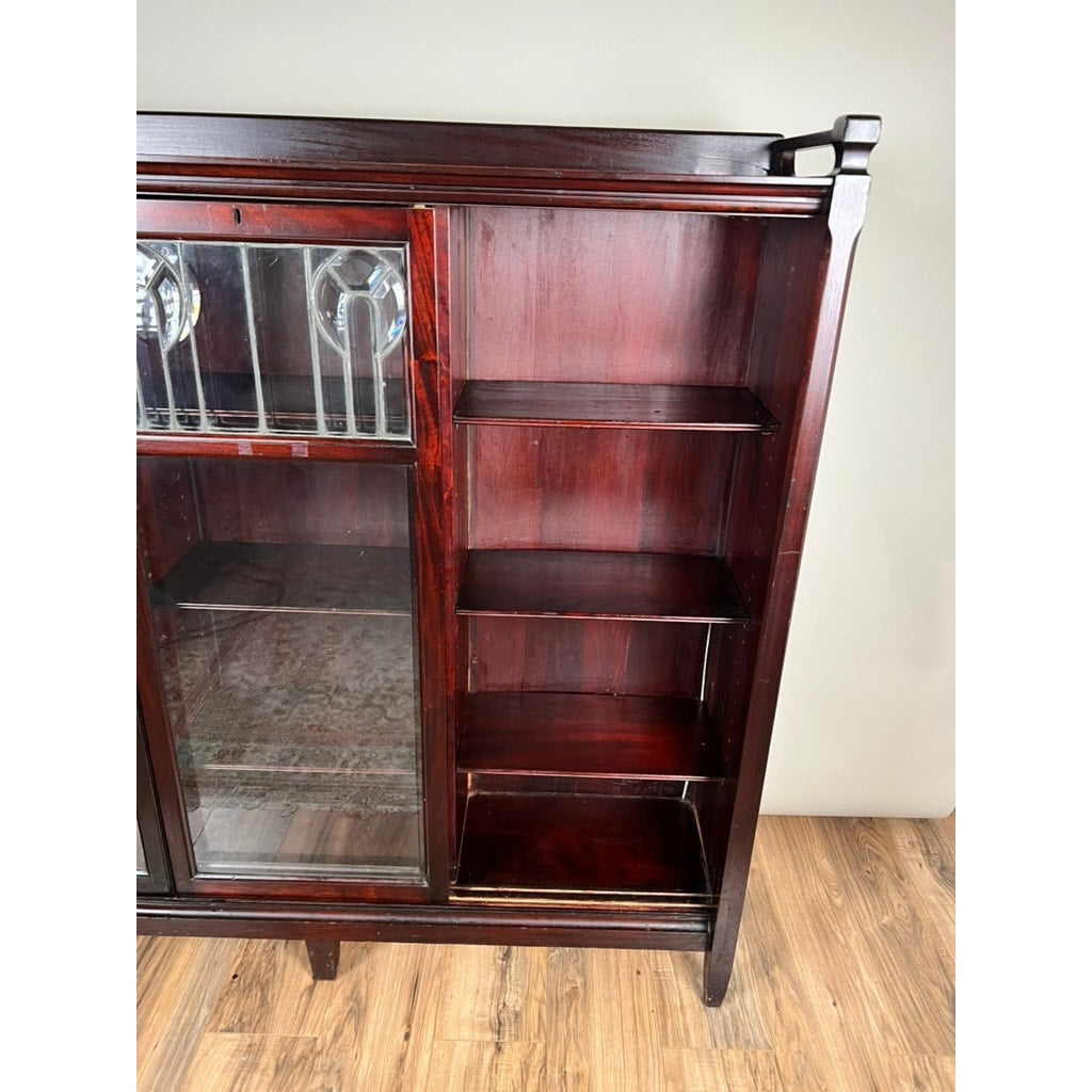 The Antique Arts & Crafts Leaded Glass Mahogany Bookcase c. 1920 stands on a wood floor against a light-colored wall, featuring leaded glass doors on the left and four open shelves on the right.