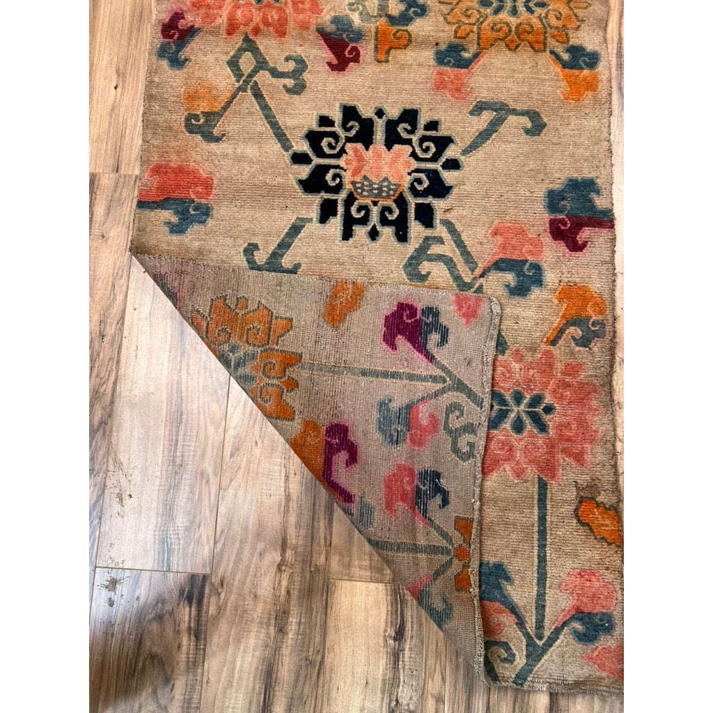 An Antique Tibetan Khaden Rug (c. Early 20th Century, 2’10” x 5’4”) with geometric orange, blue, pink, and black patterns is displayed on a wooden floor, with one corner folded to reveal its Himalayan weaving underside.