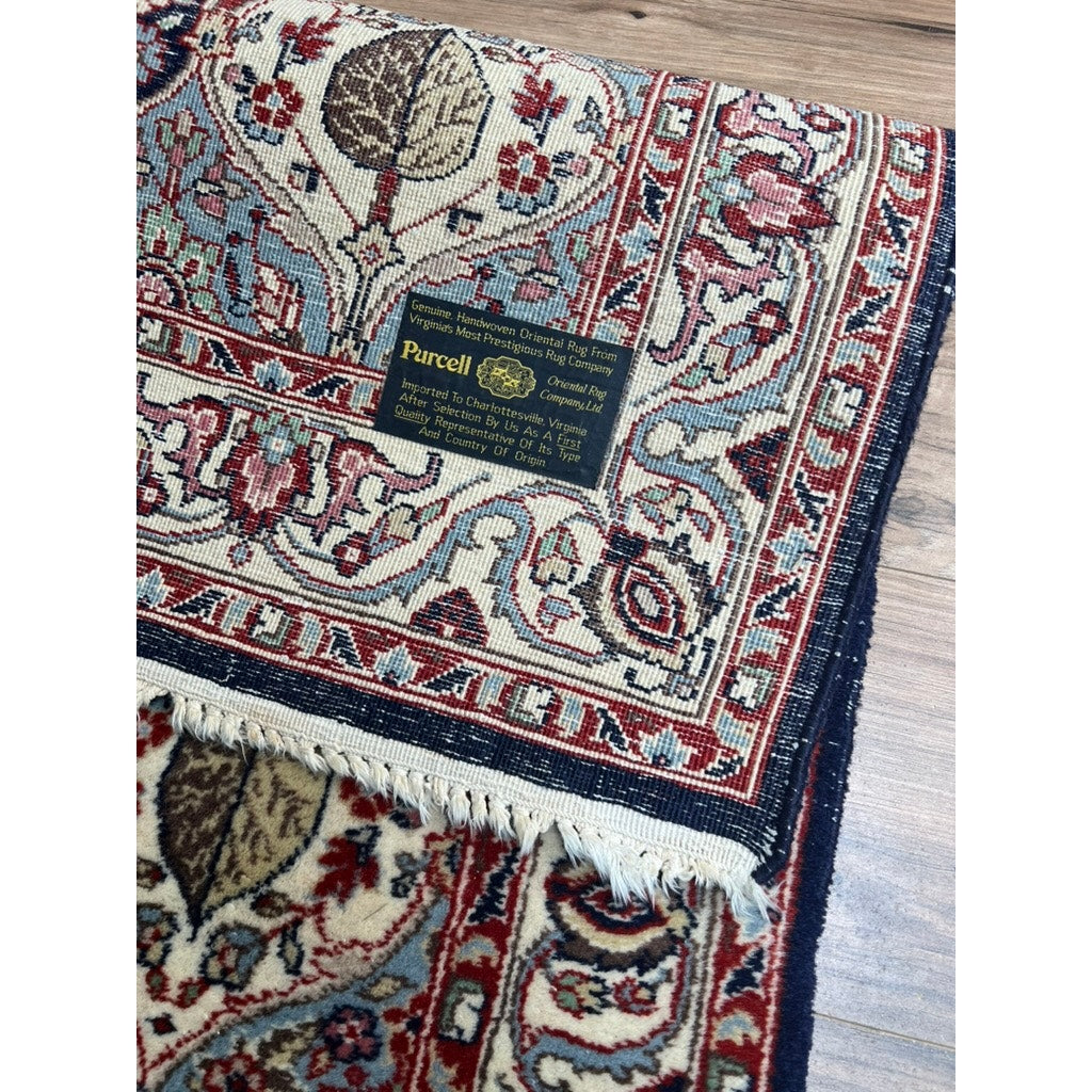 A close-up of a Vintage Hand Tied Persian Wool Rug (3’ x 5’2) with intricate red, blue, and beige patterns. The partially folded rug lays on a wooden floor, showing fringed edges; brand is unknown.