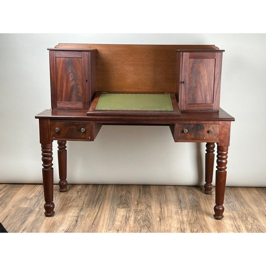 An Antique Sheraton Mahogany Plantation Secretary from the mid-19th century by Pearl City Antiques, featuring two small cabinets on the back, a central green writing surface, two front drawers, and turned legs, is displayed on a wooden floor against a plain background.