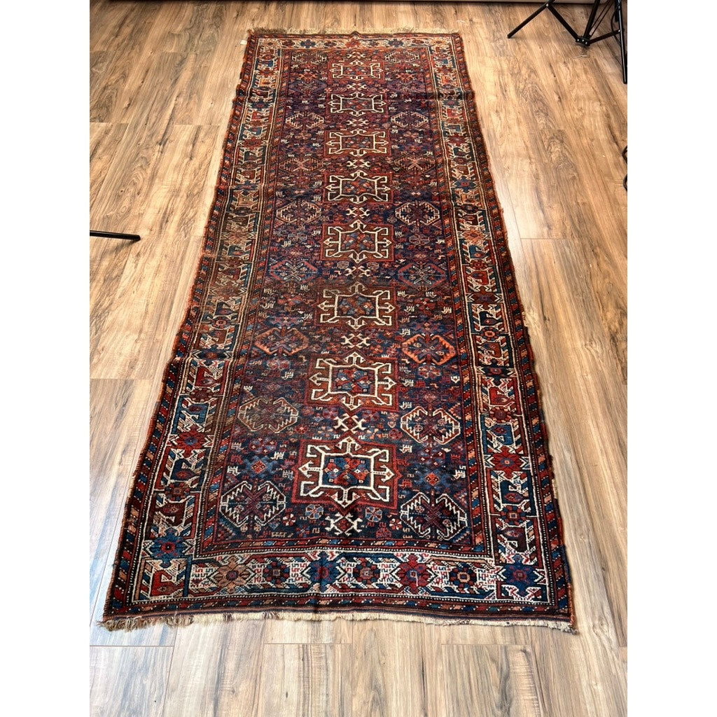 An Antique Southwest Persian Khamseh Rug (c. Late-19th Century, 3’7” x 9’1”) with geometric and floral patterns in red, blue, ivory, and brown rests on a wooden floor; edges are slightly worn with visible fringe at one end.