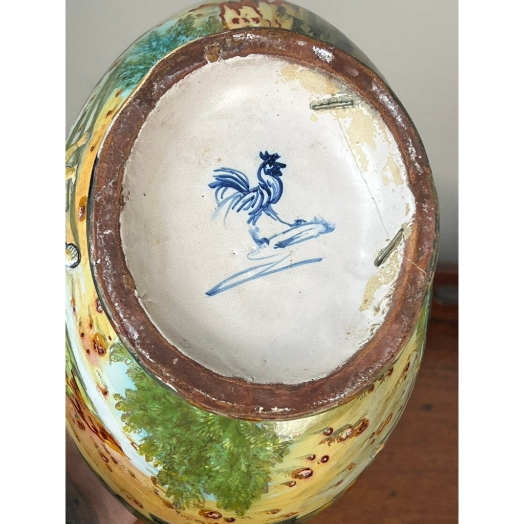 The base of this antique Italian Renaissance Revival maiolica vase (c. 1920-1940) shows an unglazed rim with typical wear and discoloration, while the side features a hand-painted decorative outdoor scene and unique winged putti handles.