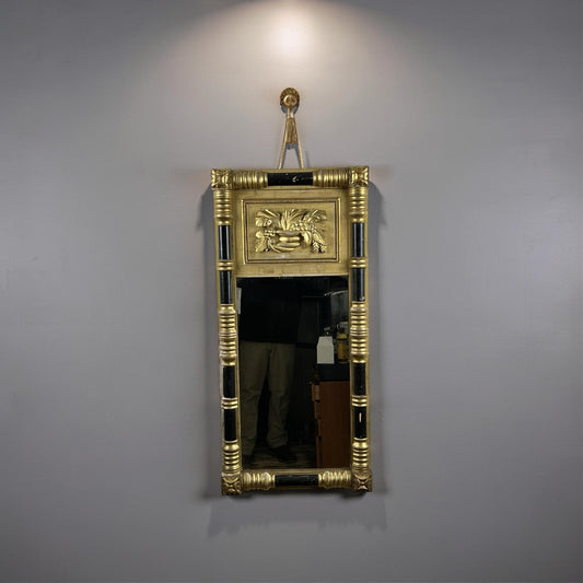 Introducing the Antique Federal Period Giltwood Mirror with Rosette Corners and Cornucopia c. 1830 from Pearl City Antiques: This rectangular wall mirror features an elegant gold and black ornate giltwood frame, a decorative top panel adorned with a sphinx, and a small round ornament above. Reminiscent of neoclassical decorative arts, this Federal period antique beautifully reflects its surroundings, including glimpses of people and furniture.