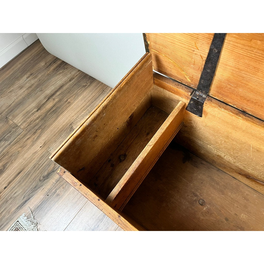 Antique 18th Century grain-painted pine six-board blanket box in mustard with till, shown open on a hardwood floor by a white wall. The chest’s natural wood grain and metal hinges are visible; part of a rug appears in the lower left corner.