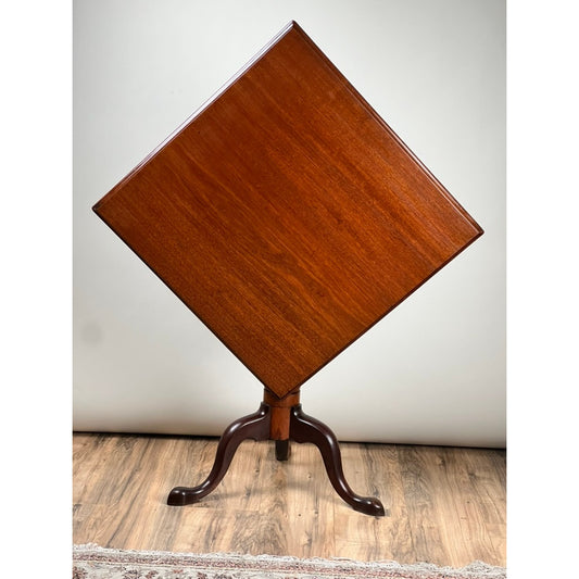 The Antique Queen Anne Mahogany Tilt Top Breakfast Table by Pearl City Antiques, dating from the late 18th century, features a square top displayed diagonally, supported by a central pedestal with three curved legs. It stands on a wooden floor against a plain background.
