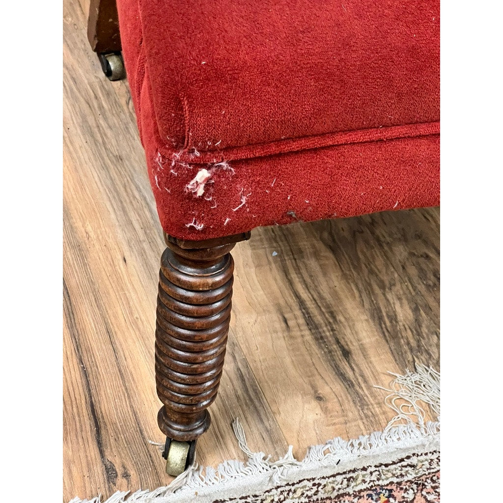 Close-up of an Antique 19th Century Wingback Chair with Rare Ring Turned Legs, featuring red velvet upholstery, a damaged frayed corner with exposed stuffing, and a carved wooden leg on caster next to a fringed rug on the wood floor.