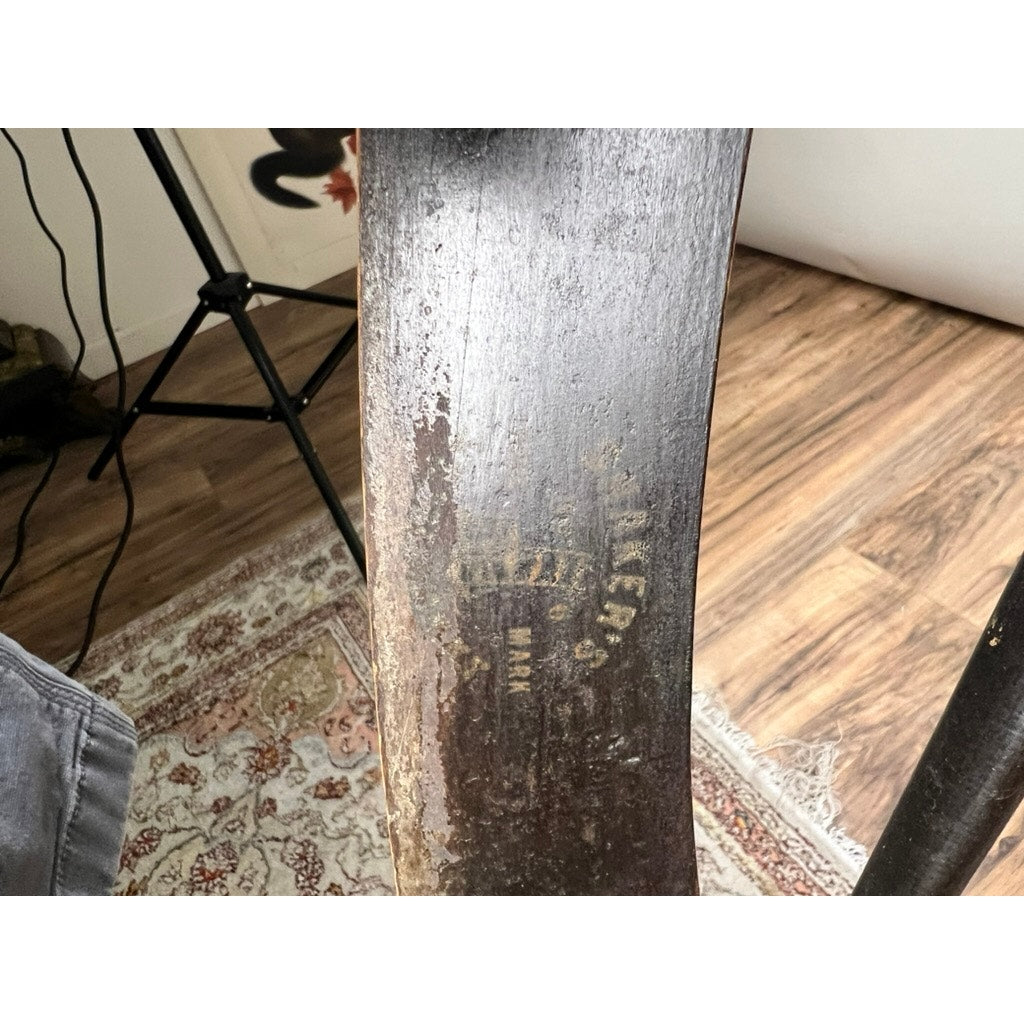 Close-up of a worn metal axe blade with faded engraving on a patterned rug. In the background is an Antique Shaker #6 Tape Seat Rocker by Mt. Lebanon Shakers, late 19th century, plus part of a tripod and wooden floor. Brand: Unknown.