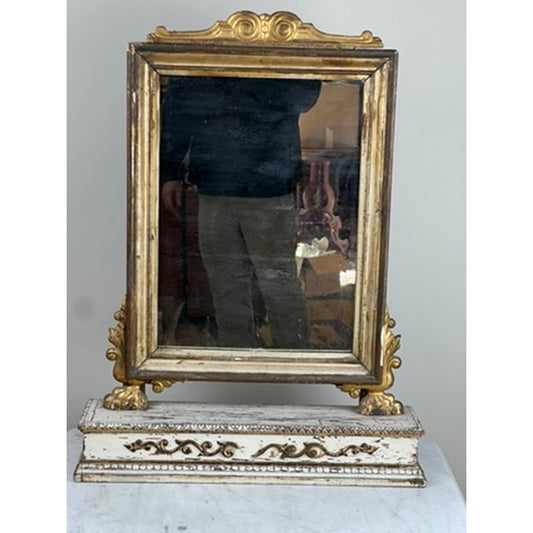 An Antique French Empire Gilt Wood Vanity or Table Mirror by Pearl City Antiques, dating to the mid-19th century, stands on a decorative base with an ornate gold and white frame. Echoing the refined style of French Empire design, its gilt wood detailing brings an air of antique elegance. The mirror reflects part of a person and surrounding furniture, capturing timeless sophistication.
