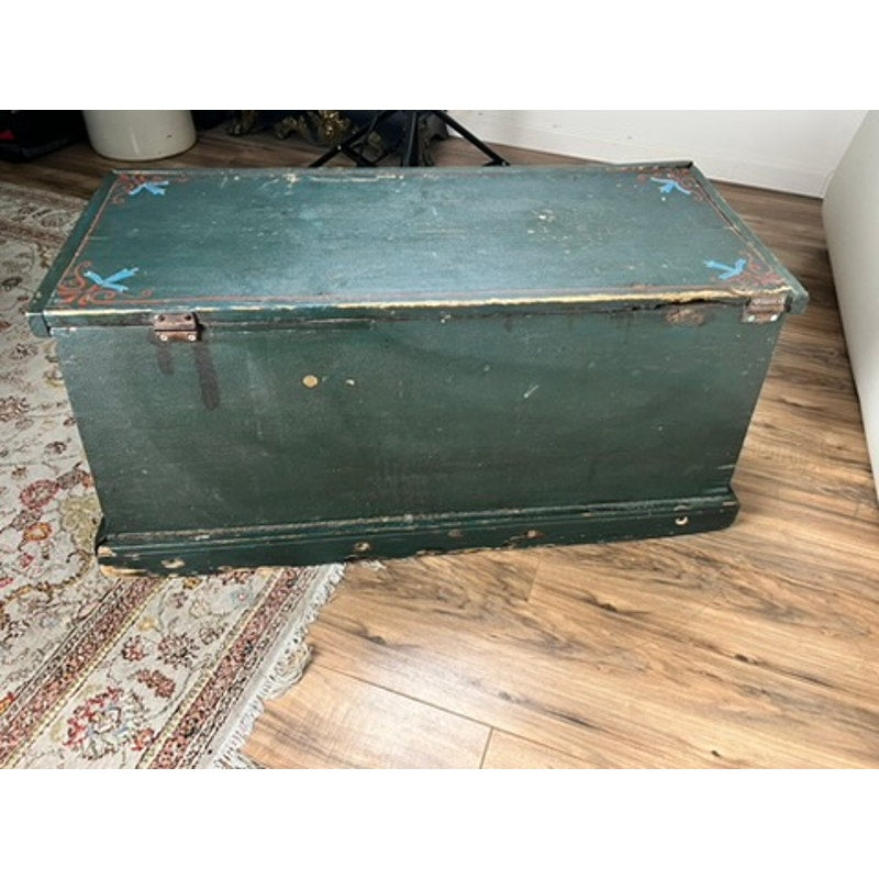 An Antique Paint Decorated English Sea Chest from the late 19th century, showing signs of wear and age, features dark green paint, metal hinges, and faint patterns. It rests on a wooden floor beside a patterned rug. Brand is unknown.