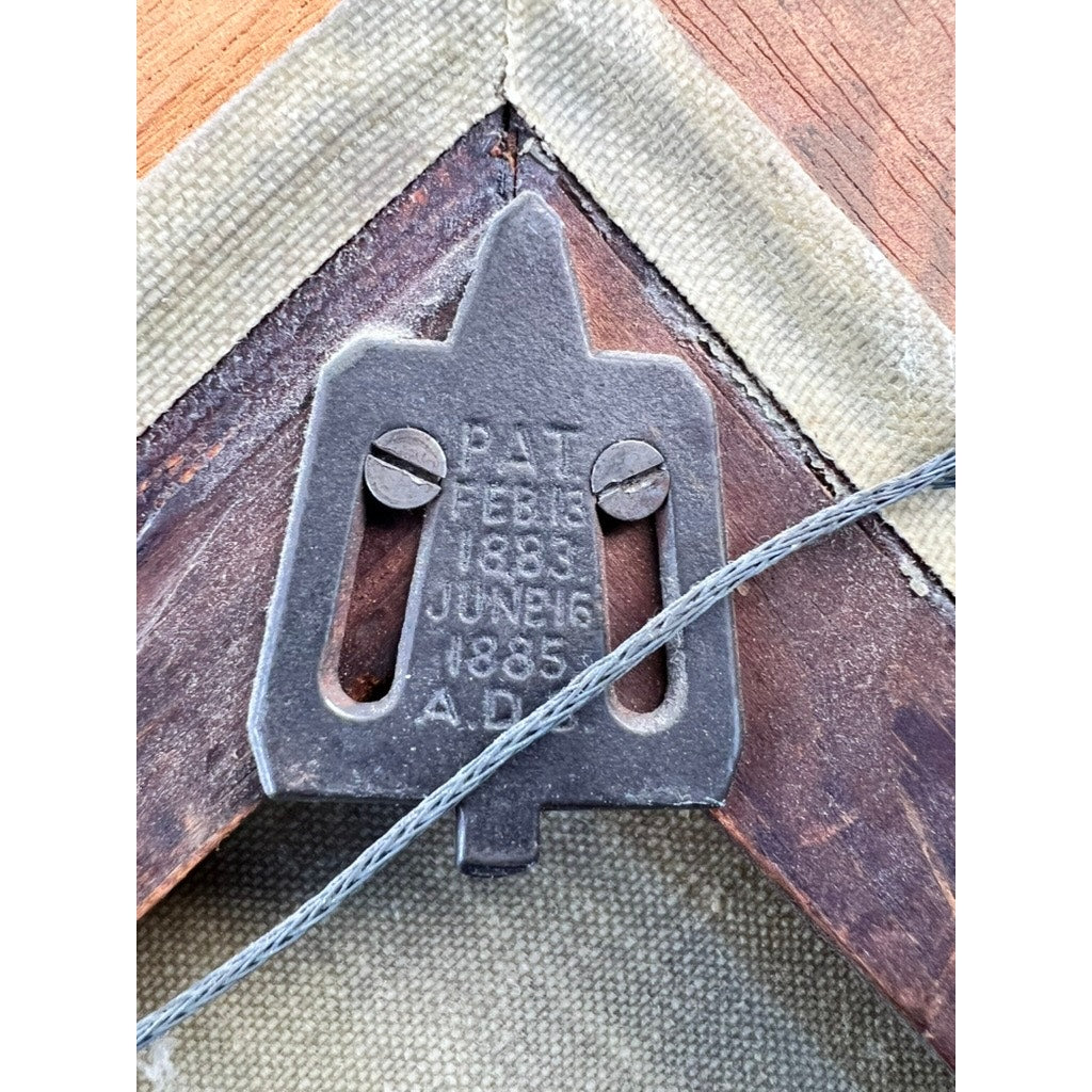 Close-up of a metal picture hanger attached to the back of an Antique American School Farmstead Landscape Oil Painting, Circa 1900, with two screws and engraved patent dates: Feb 13, 1883 and June 16, 1885. A wire is looped through the hanger.