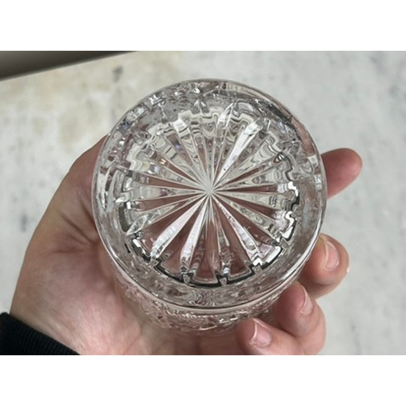 A hand holds a Pair Fostoria Stowe Double Old Fashioned Tumbler Glasses upside down, showing the decorative starburst cut glass pattern on the bottom against a light, textured background.