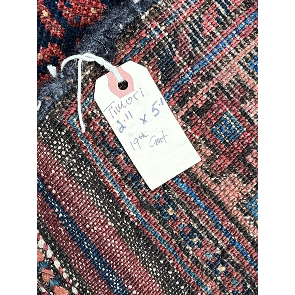 A close-up of an Antique Timuri Baluch Rug from Western Afghanistan, c. 19th Century (2’11 x 5’1), showing intricate red, blue, and brown geometric designs and a handwritten tag with the rug’s size and origin. Brand: Unknown.