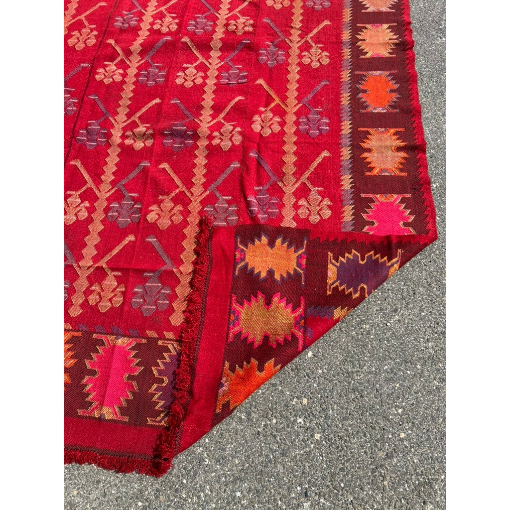The Vintage Handmade Bulgarian Thracian Kilim Flat Weave Rug c. Early 20th Century (5’4 x 11’7) features vibrant red geometric patterns in gold, orange, and brown and is shown on a gray surface with one corner folded to reveal the underside.