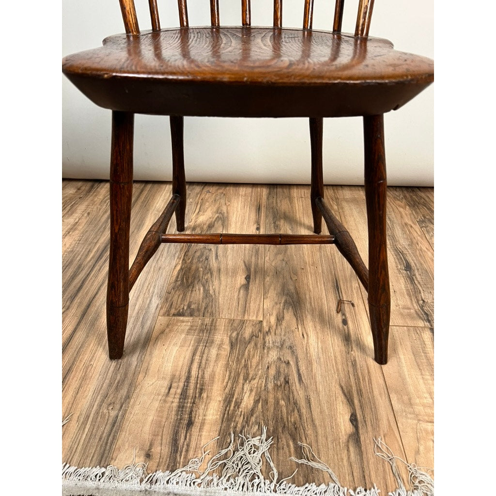 A close-up of an Antique Step Down Windsor Chair in Oak and Pine (early 19th century) shows its curved seat, spindle back, legs, and base on a wood-patterned floor with part of a fringed rug also in view. Brand: None/Unknown.