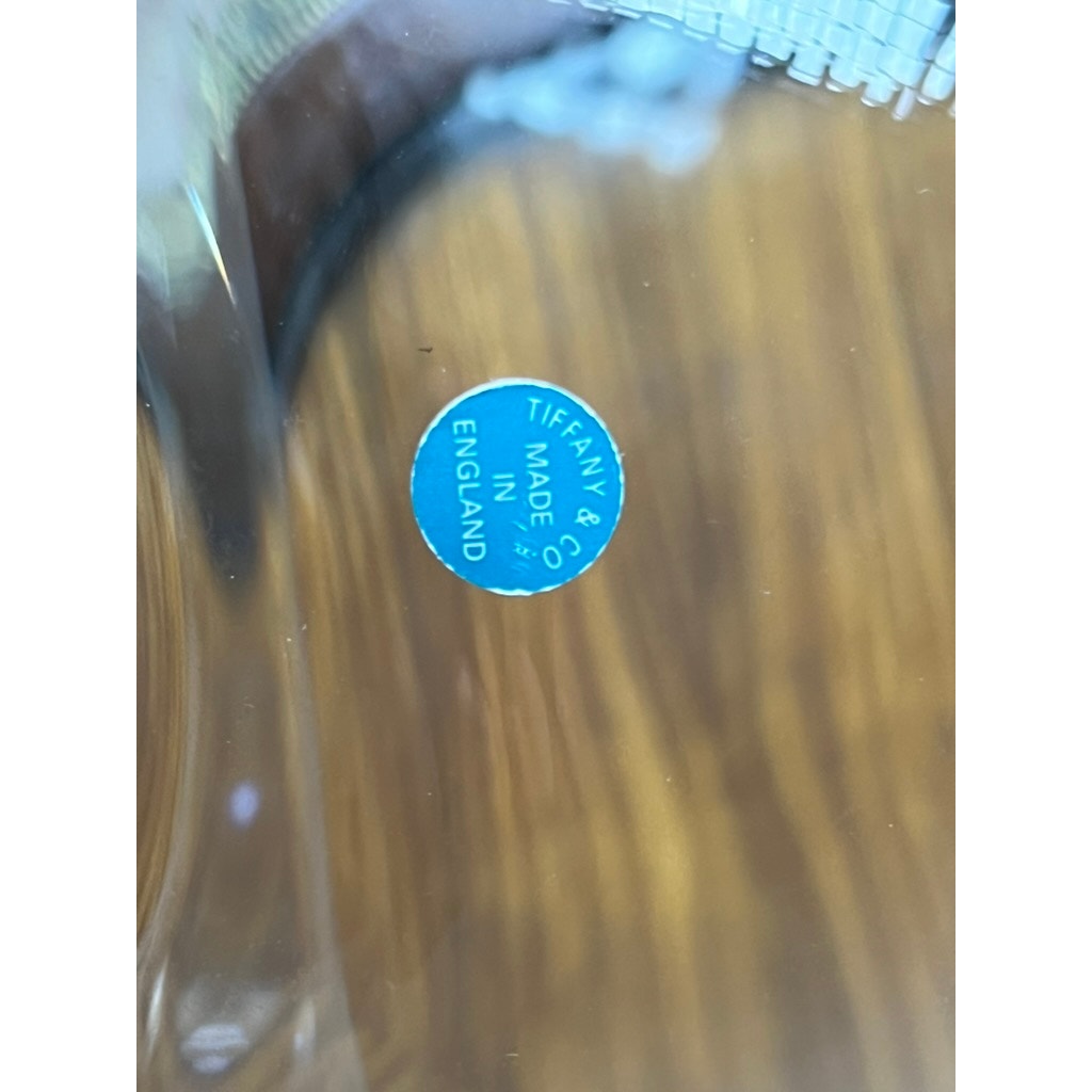 Close-up of a small blue sticker reading "Tiffany & Co. Made in England" on a Vintage Tiffany & Co. 8” Crystal Revere Bowl, displayed on a wooden table.