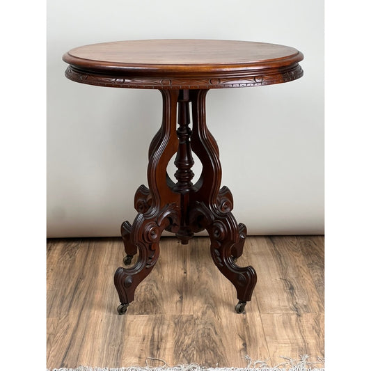 The Antique Victorian Walnut Lamp Stand c. Late 19th Century by Pearl City Antiques features an ornate round wooden pedestal with intricate carved details, four elegantly curved legs set on small wheels, and a rich dark polished finish. Reminiscent of classic Victorian design, this stunning 19th-century piece stands gracefully on a wood floor against a plain light-colored background.