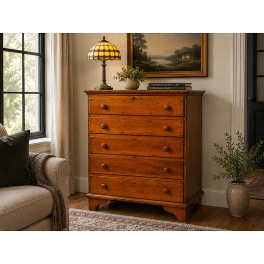 A Late 18th Century New England Pine Two-Drawer Blanket Chest by Pearl City Antiques, dating from around 1780–1810, stands against the wall in a cozy living room with a couch, potted plant, large windows, and a framed painting. The chest has a lamp and books on top, evoking the charm of early American country furniture.