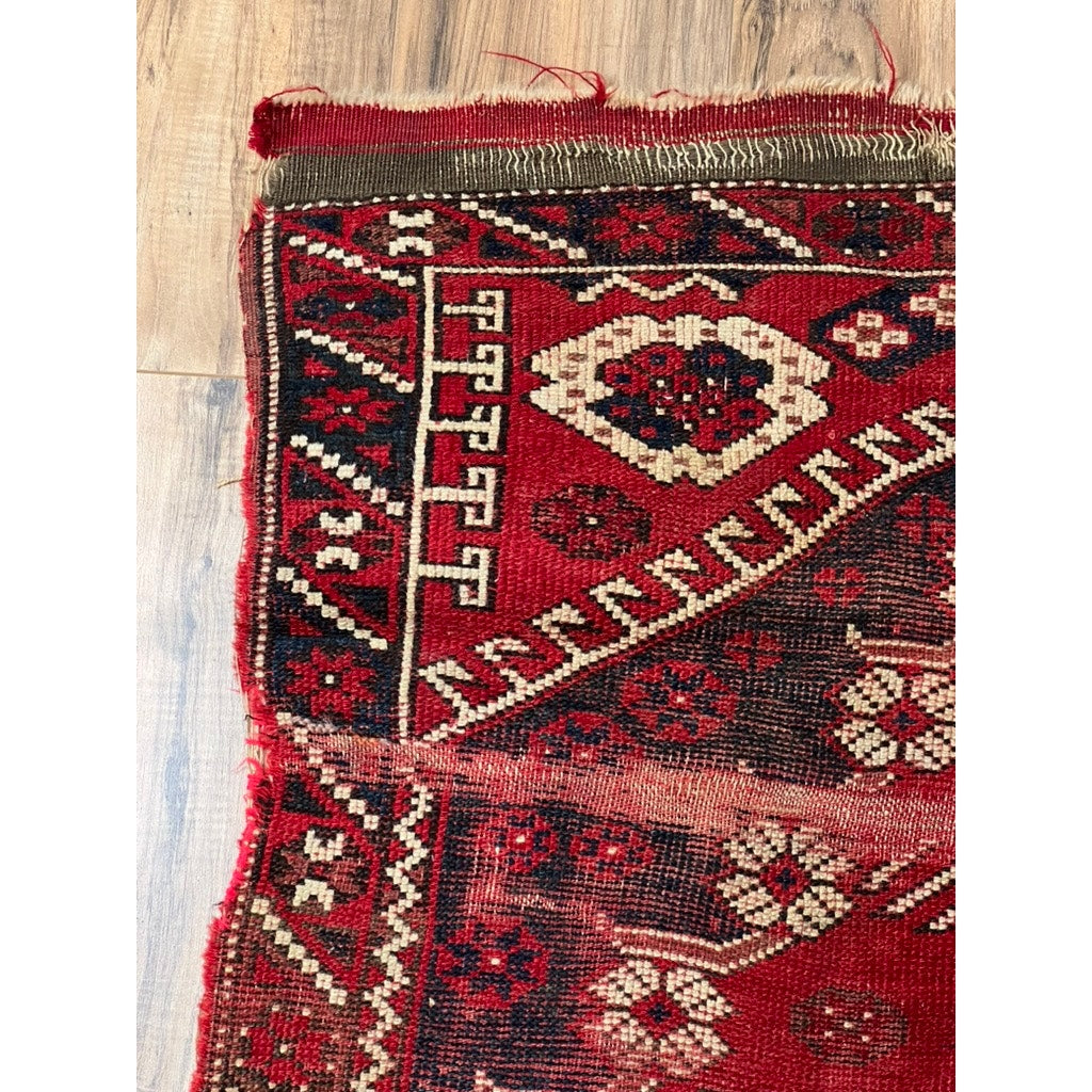 A close-up of the corner of an Antique Kazak Ruby Red Rug c. Late 19th Century (3’1 x 4’4), featuring geometric and floral motifs, with frayed edges and visible wear visible on a wooden floor. Brand: Unknown.