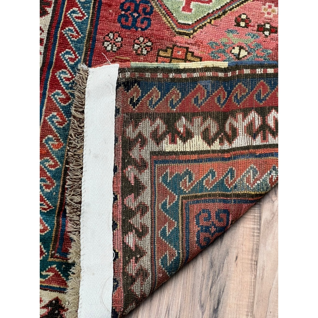 The Antique Caucasian Kazak Rug c. Late 19th Century (2’10 x 3’11) features vibrant geometric and floral patterns, is partially folded to show its underside, and lies on a light wooden floor with a white edge along the fold.