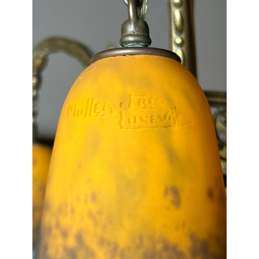 Close-up of a yellow and orange glass shade etched "Muller Frères Lunéville," attached to the brass fixture of a Vintage Muller Frères Lunéville French Art Glass Four Light Chandelier, with a decorative background.