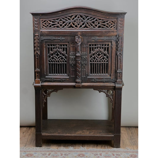 The Antique 19th Century Belgian Gothic Revival Court Cupboard features intricate Gothic-style carvings, arched door panels, decorative patterns, and stands on four legs with an open lower shelf. Placed on a wooden floor near a patterned rug.