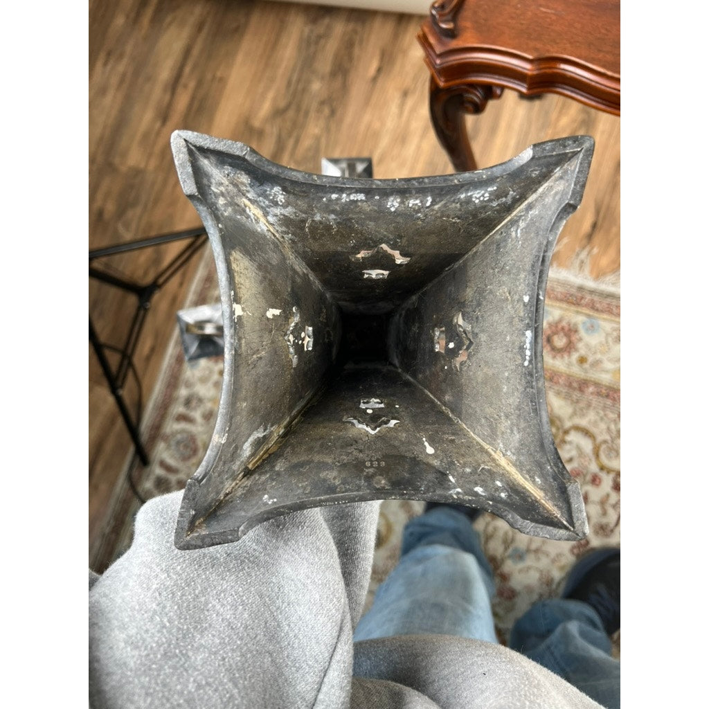 View from above of a person holding a weathered, square metal vase with a chipped interior; wooden floor, patterned rug, furniture, and an Antique Art Nouveau Five Arm Candelabra by Poole Silver seen in the background.