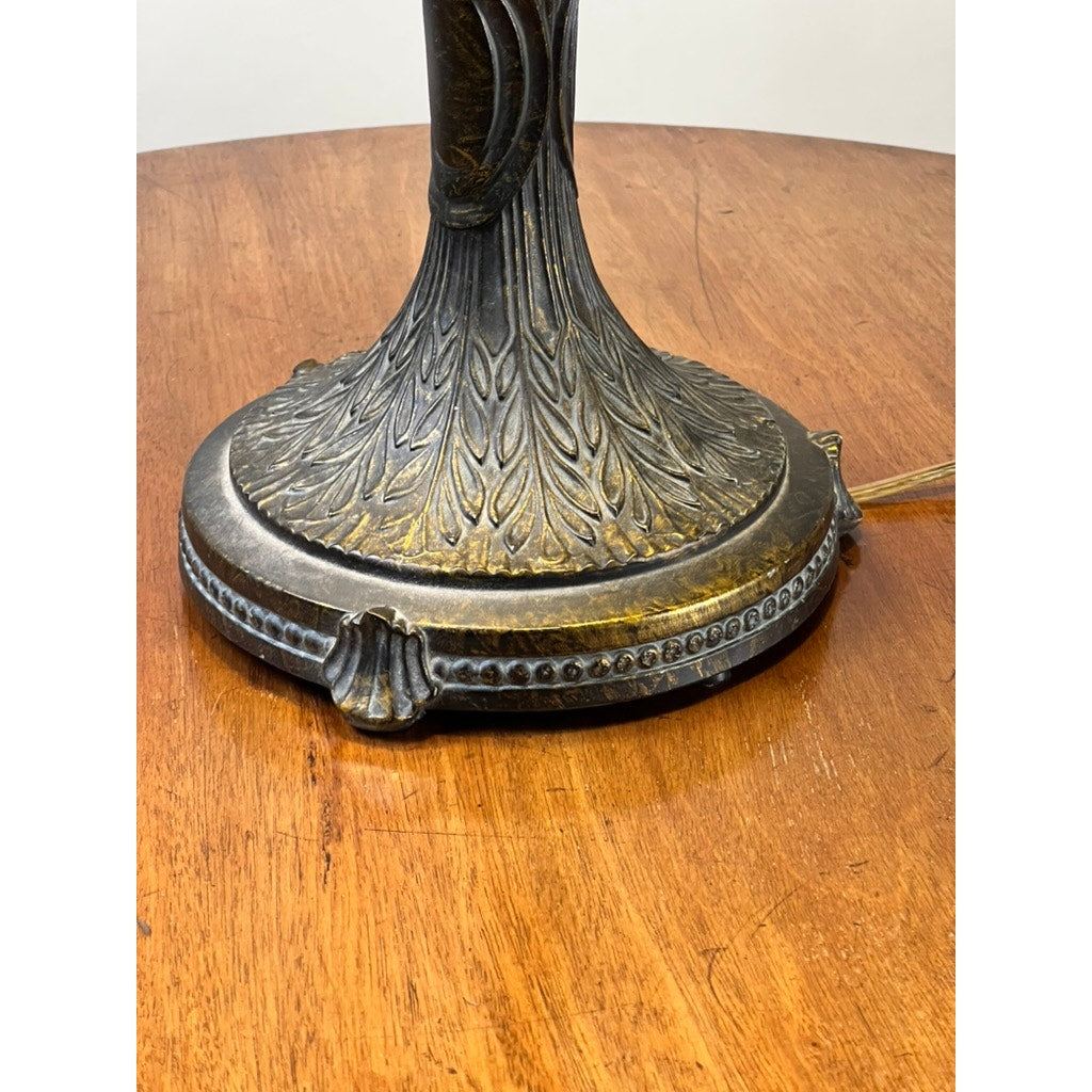 Close-up of the ornate metal base from a Vintage Tiffany Style Floral Stained Glass Table Lamp, featuring leaf patterns and resting on polished wood. A power cord extends from the base to the right. Brand: None/Unknown.