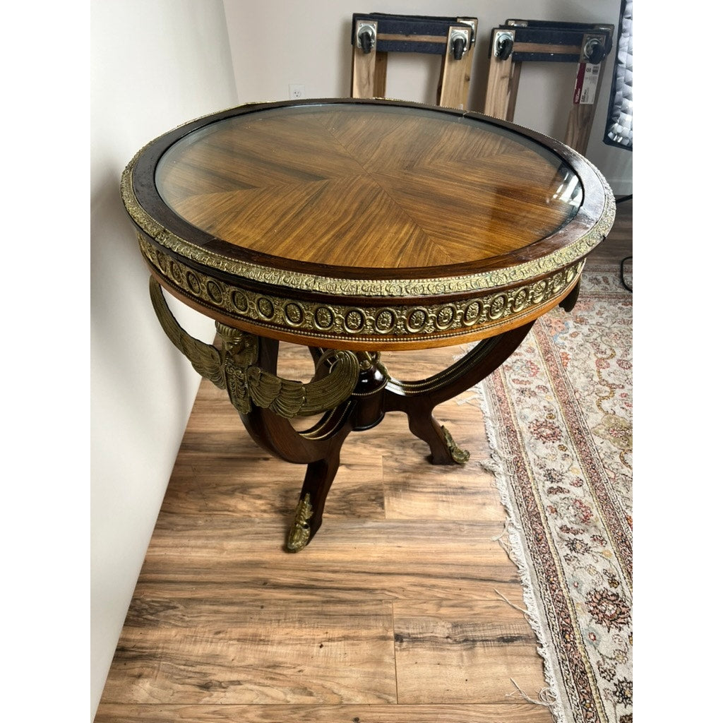 An Antique French Empire Revival Gueridon Table with ormolu mountings (early 20th century) stands on a wood floor near a patterned rug and plain white wall, featuring elegant kingwood veneer, glass top, and ornate gilt bronze trim.