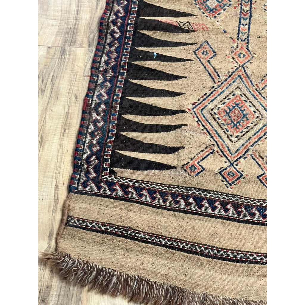 A close-up of the Antique Baluch Sofreh Runner c. Mid 19th Century (2’3 x 9’2) highlights tribal geometric patterns in red, black, and blue on beige with fringed edges, displayed on a wooden floor. Brand: Unknown.