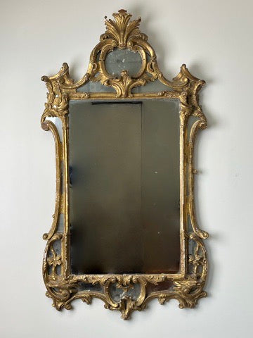 The Antique Late 18th Century Italian Baroque Gilt Gesso Wall Mirror with Mirrored Openwork by Pearl City Antiques, with elaborate gold scrollwork and crest, elegantly adorns a plain light-colored wall.