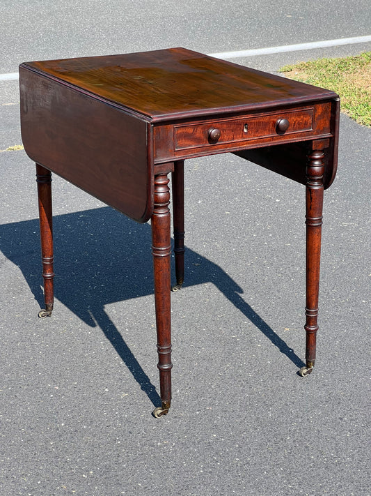 The Antique Mahogany George III Sheraton Drop Leaf Table c. Late 18th Century by Pearl City Antiques stands on asphalt near grass; it features turned legs with casters and a central drawer with round knobs—an elegant period piece.