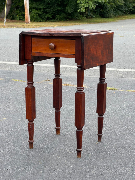 The Antique New York Sheraton One Drawer Stand c. 1800 by Pearl City Antiques, featuring four turned legs, a single round-knob drawer, and drop-leaf sides, is displayed outdoors on asphalt with trees and a utility pole in the background.