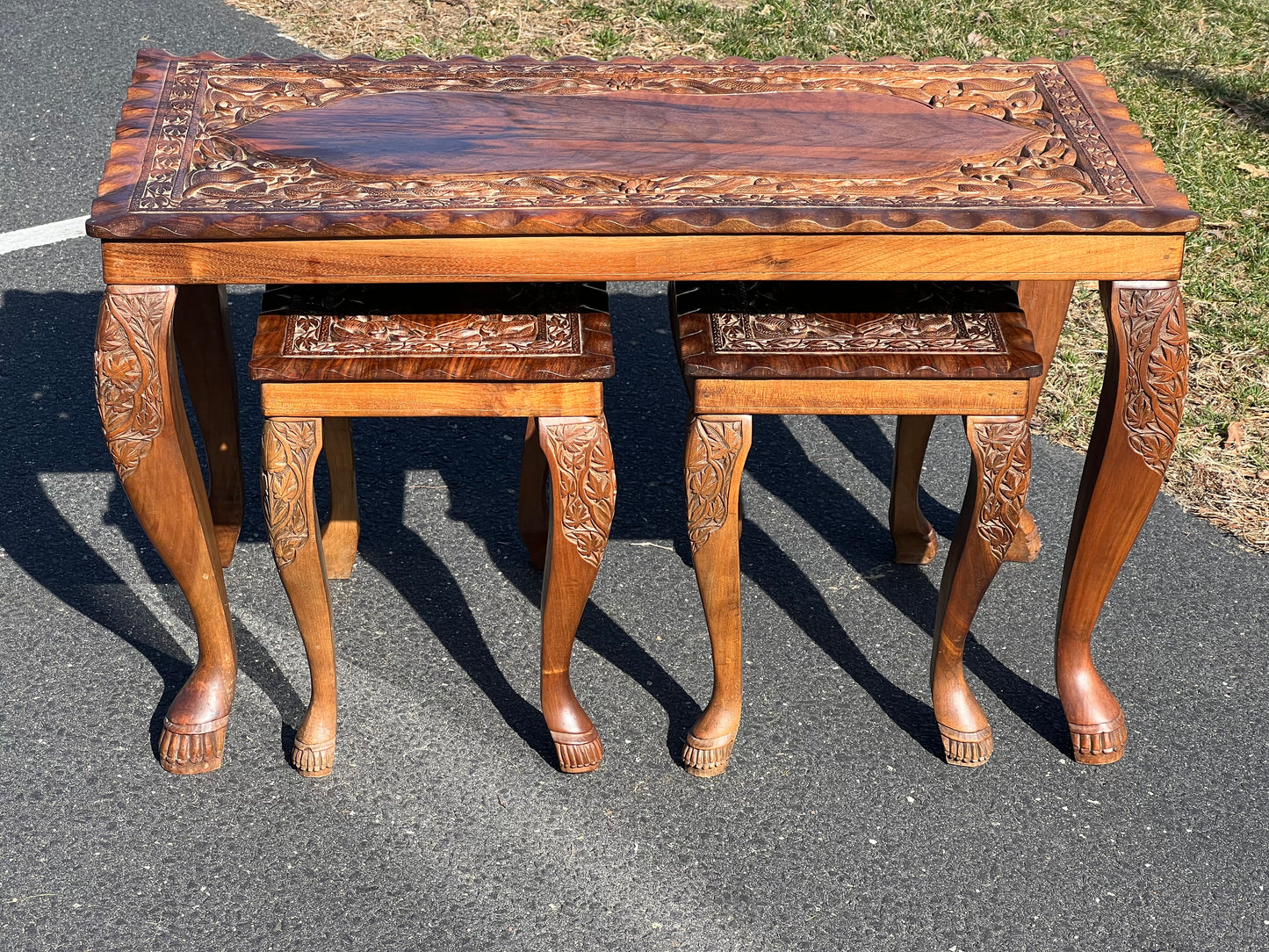 The Pearl City Antiques Vintage Hand Carved Asian Fruitwood Nesting Tables feature detailed floral carvings and curved legs with paw-shaped feet, including two smaller tables, all crafted in wood and displayed outdoors on asphalt.